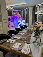 En spiseplads med et langt bord dækket til fest, med et oplyst skilt med '25', blomsterarrangementer og en lysekrone.