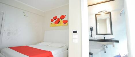 A hotel room with a bed, a red blanket, a white headboard, a wall-mounted painting, a bathroom with a sink, mirror, and a light fixture.