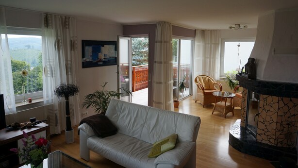 A living room with a white sofa, a fireplace, and a balcony with a view of greenery. - Like new EFH for 6 pers. with garage in close proximity to Sorpesee (Langscheid)