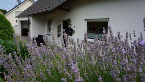 A white house with a tiled roof, a front porch, and a garden with lavender flowers. - Like new EFH for 6 pers. with garage in close proximity to Sorpesee (Langscheid)