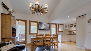Un salón espacioso con un sofá oscuro, una mesa de comedor de madera y sillas. Un candelabro único cuelga del techo, y un televisor está montado en la pared.