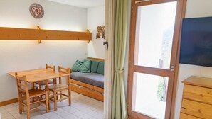Une pièce avec une table et des chaises en bois, un canapé avec des coussins, un téléviseur fixé au mur et une fenêtre avec des rideaux.