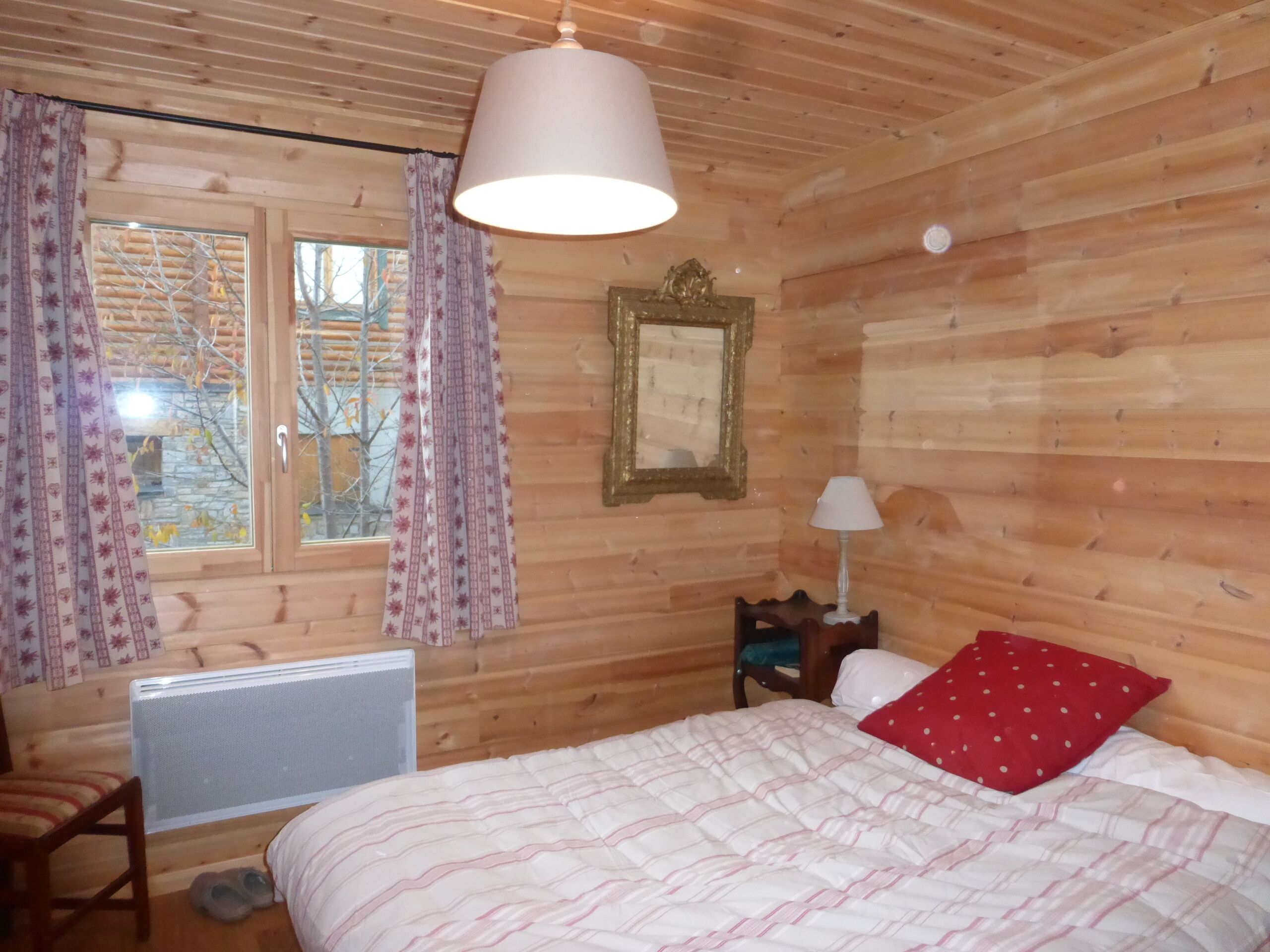 A wooden cabin room with a bed, a chair, a radiator, and a window with curtains.
