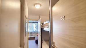 Un couloir étroit avec un lit superposé et une fenêtre. La pièce a une porte en bois portant le numéro 323.