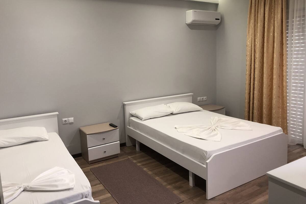 A hotel room with two beds, a bedside table, a nightstand, a wall-mounted air conditioner, and a window with curtains.