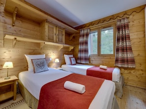 A wooden cabin room with two beds, a window with curtains, and a lamp on a bedside table.