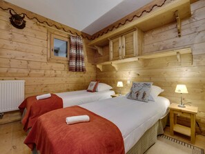 Ein Zimmer in einer Holzhütte mit zwei Betten, einem Vorhangfenster, einem Heizkörper und einem kleinen Nachttisch aus Holz mit einer Lampe.