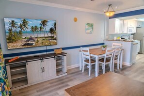 Une cuisine avec des meubles blancs, une table à manger en bois avec des chaises blanches, un téléviseur fixé au mur et un petit îlot central doté d’un évier et d’un plan de travail.