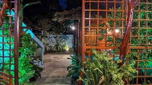 Un sendero exterior bien iluminado que conduce a una estructura de madera con una cerca tipo celosía, rodeada de abundante vegetación y plantas.