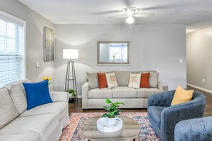 Un salón con un sofá color beige, un sillón azul, un sillón gris, una mesa de centro con una planta, una lámpara de pie y un ventilador de techo.