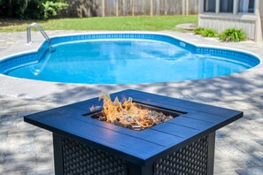 Un foyer extérieur rectangulaire, avec une flamme visible, installé sur une terrasse avec une piscine en arrière-plan.