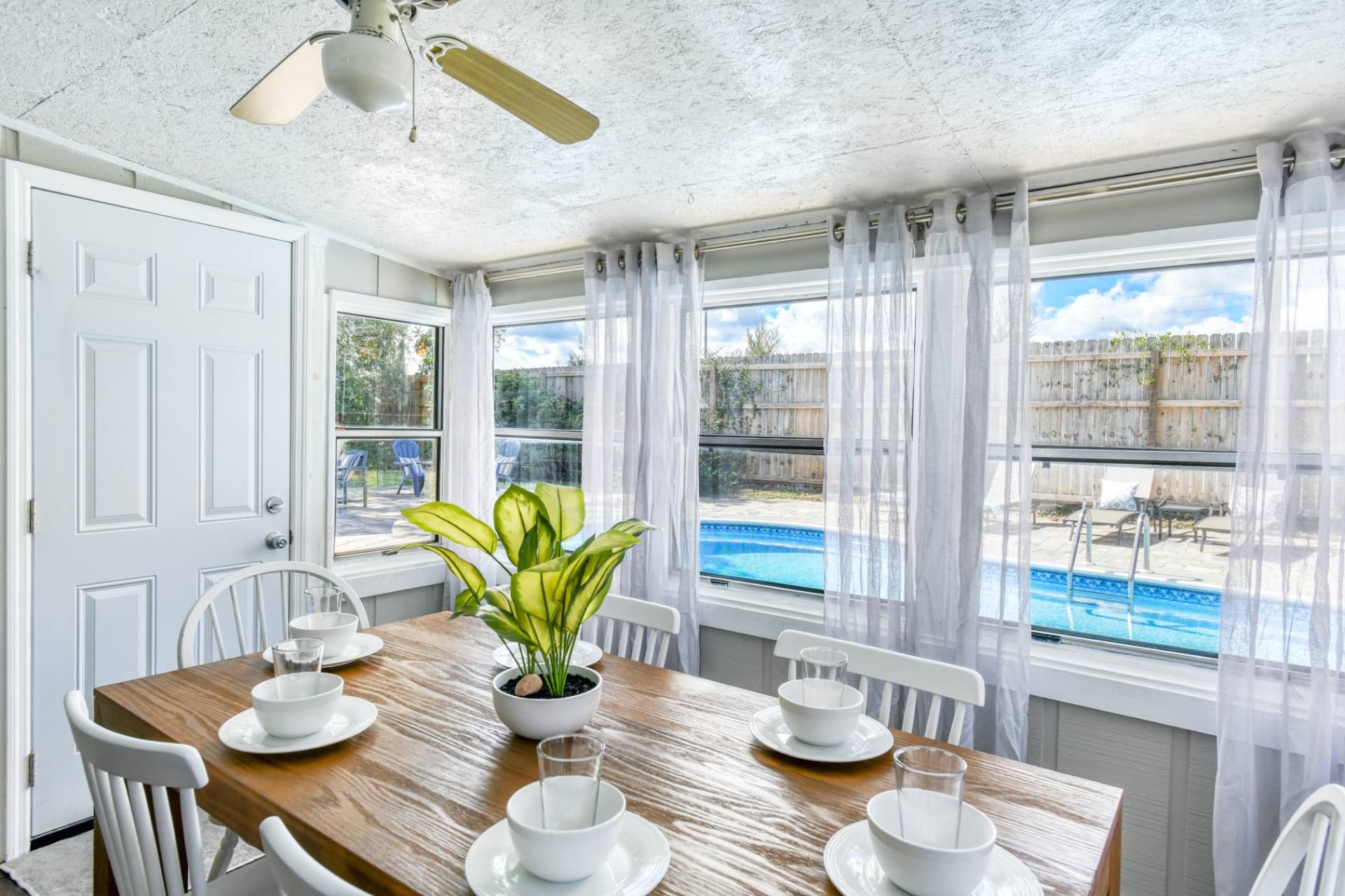 A dining area with a wooden table set for four, white chairs, a potted plant, and a view of a swimming pool through large windows.