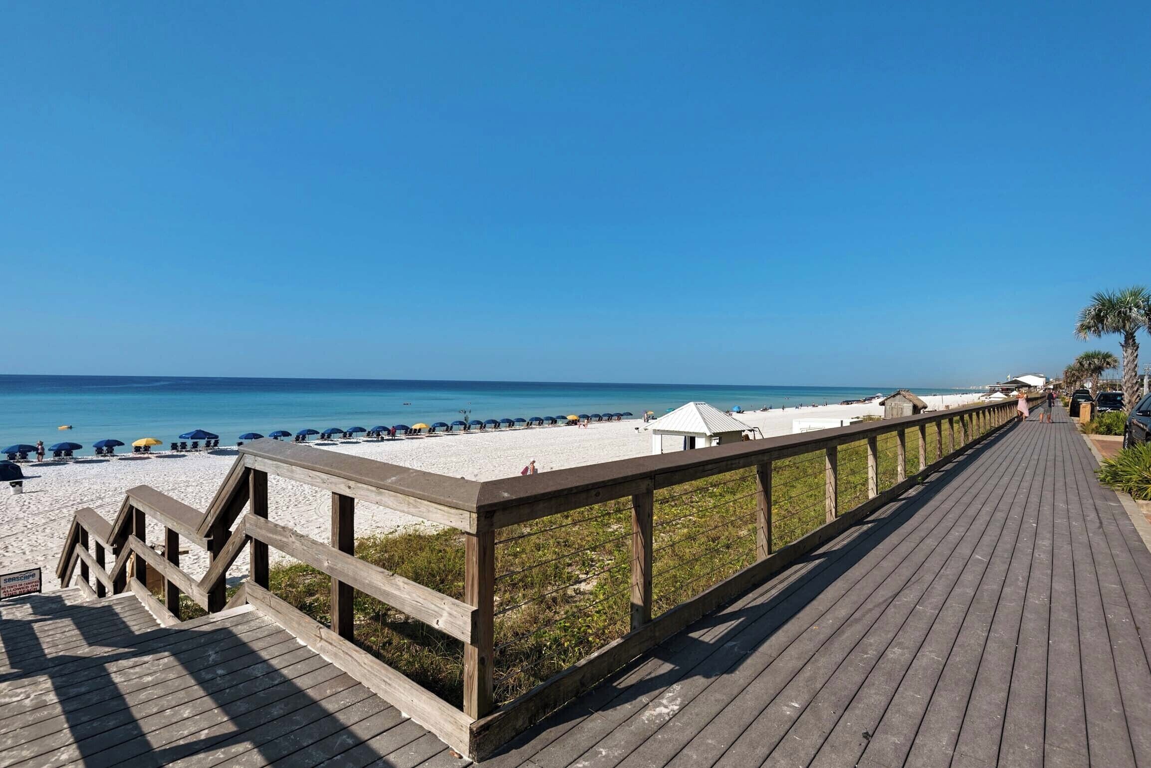 A wooden boardwalk leading to a beach with umbrellas and a small structure.