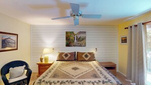 A bedroom with a bed, a ceiling fan, a nightstand with a lamp, a chair, and a window with curtains. - Driftwood 4 bedrooms, 3 bathrooms (Jekyll Island)
