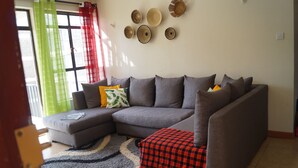 Un salon avec un canapé d’angle gris, un tapis à motifs et un pouf à carreaux rouges et noirs.