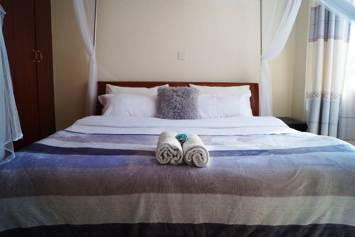 A neatly made bed with a striped bedspread, a pair of rolled towels, and a wooden headboard.