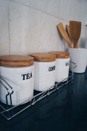 Tres recipients de ceràmica blanca amb tapes de fusta, etiquetats com a “TEA”, “COFFEE” i “SUGAR”, sobre una estructura metàl·lica davant d'una paret de rajola.