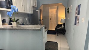 A compact kitchen with a microwave, cabinets, and a sink. There are two stools and a small table with a lamp. A door leads to a living area with a couch and a coffee table. - Kozy Bahamas Getaway (Nassau)