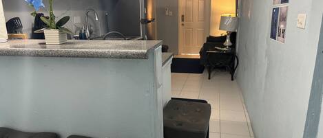 A compact kitchen with a microwave, cabinets, and a sink. There are two stools and a small table with a lamp. A door leads to a living area with a couch and a coffee table.
