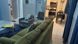 A living room with a green sofa, a coffee table, and a television. There is a kitchen area with a microwave, oven, and cabinets. A door with a wreath is visible, and a sign on the wall reads "HOME". - Kozy Bahamas Getaway (Nassau)