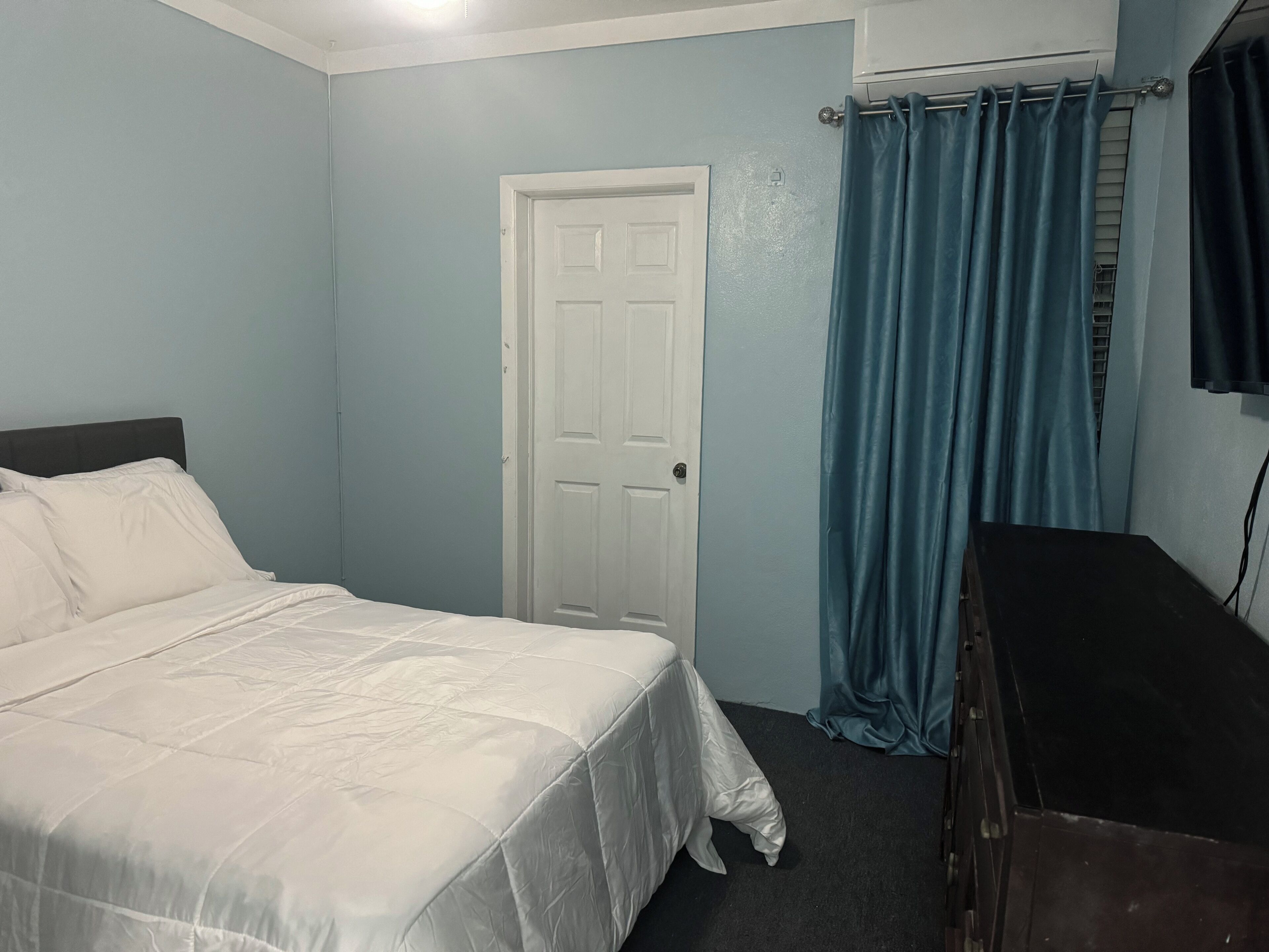 Une chambre à coucher avec un lit, une commode, une porte et une fenêtre avec des rideaux.