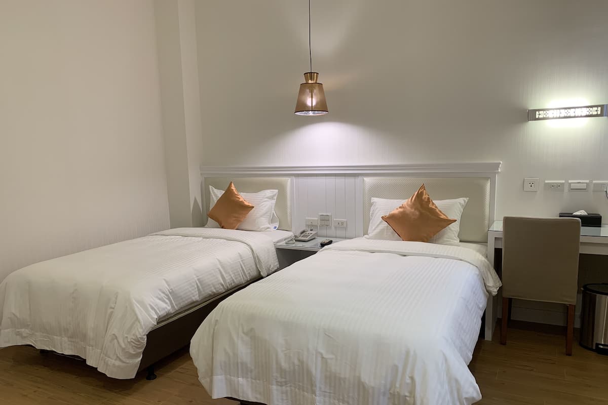 A hotel room with two single beds, a desk, a chair, and a hanging light fixture.