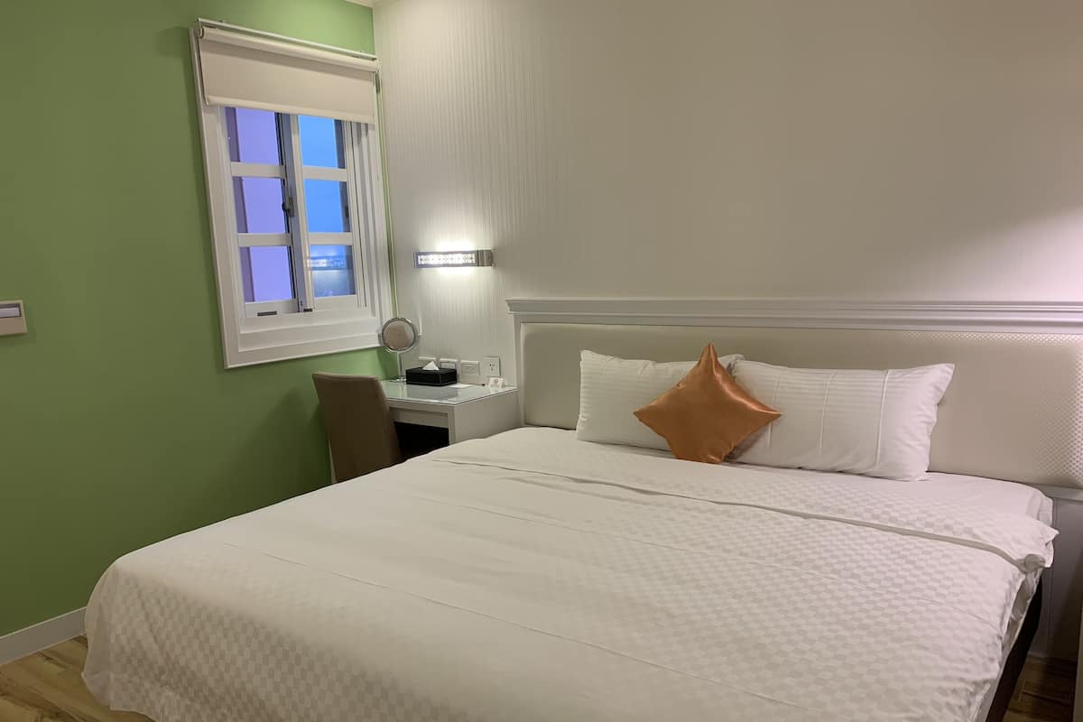 A hotel room with a bed, a desk with a chair, a window, and a wall-mounted lamp.