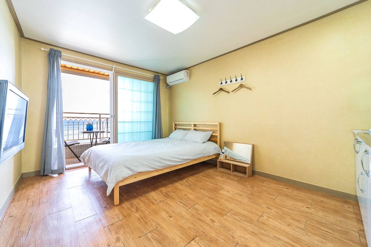 A bedroom with a wooden bed, a bedside table, a small chair, a TV, and a balcony with a view.