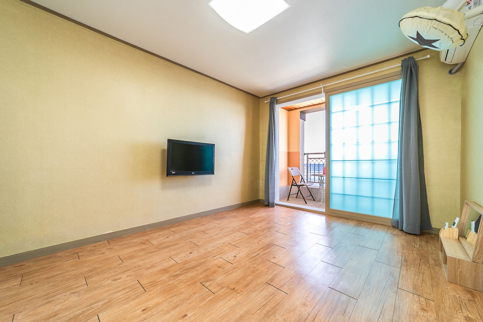 A spacious room with a large sliding glass door leading to a balcony, a flat-screen TV mounted on the wall, and a wooden floor.