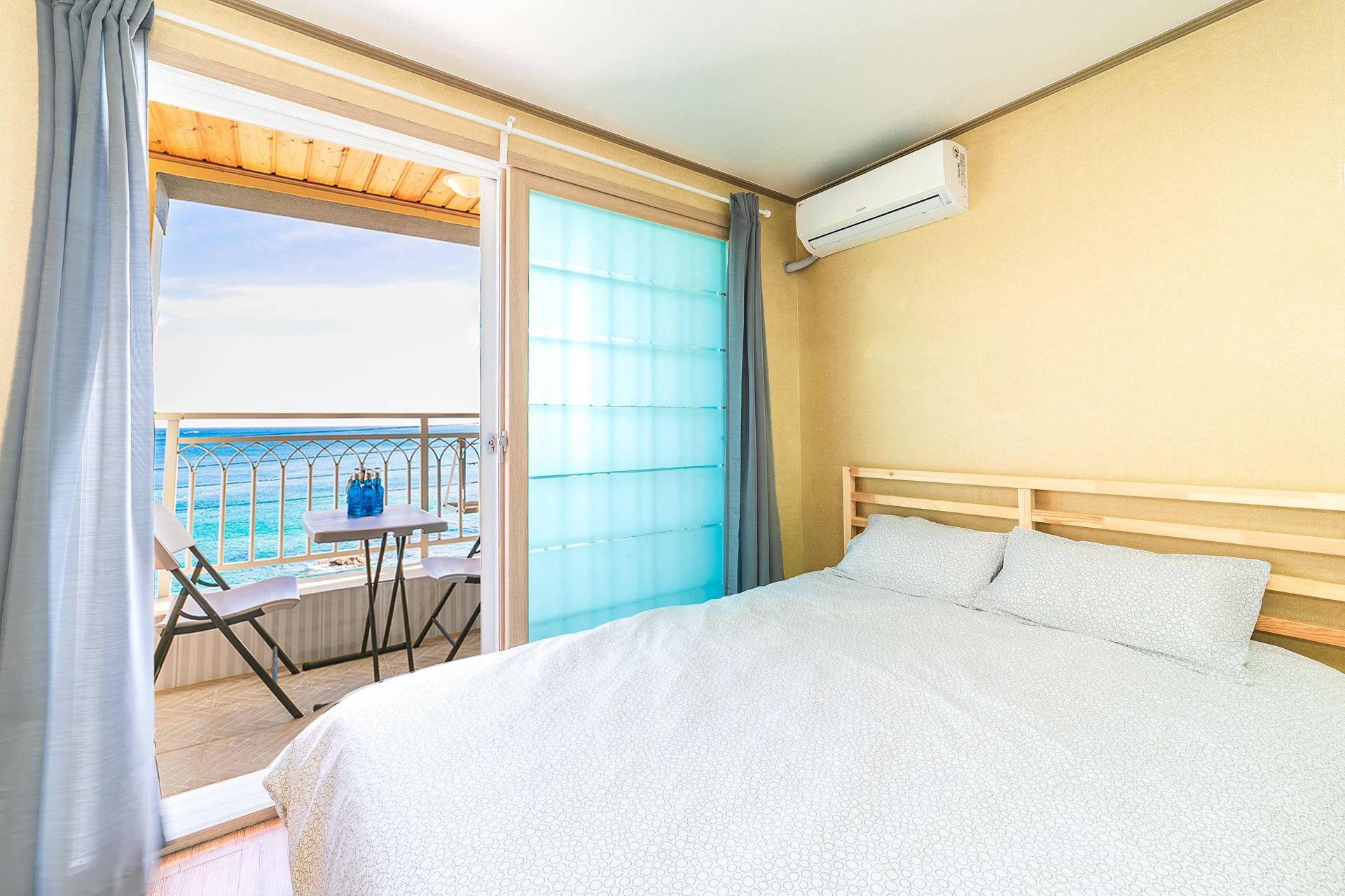 A hotel room with a bed, a table with chairs, a view of the ocean, and an air conditioning unit.