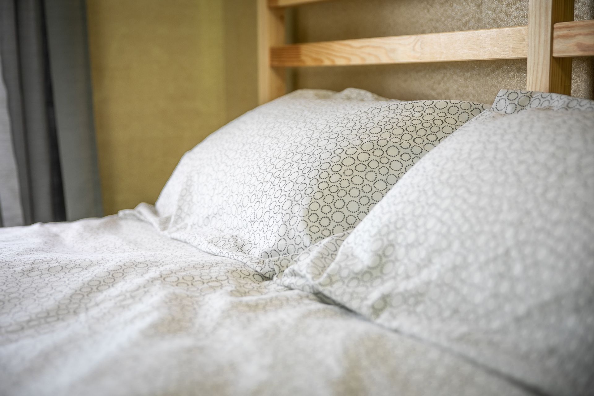 Un lit avec une couette blanche à motifs et des oreillers, une tête de lit en bois et un mur de couleur neutre.