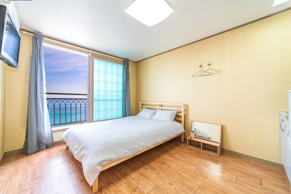 A bedroom with a bed, a small bedside table, a nightstand, a window with curtains, and a view of the ocean.