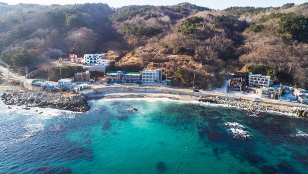 Vista aérea de un pueblo costero con edificios a lo largo de la costa, aguas turquesas cristalinas y un paisaje montañoso al fondo.