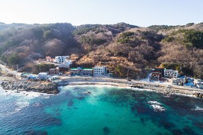 Vista aérea de un pueblo costero con edificios a lo largo de la costa, aguas turquesas cristalinas y un paisaje montañoso al fondo.