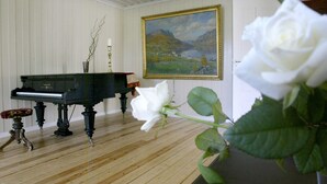 A room with a grand piano, a framed landscape painting, and a white rose. - Sommerhotellet (Vang)