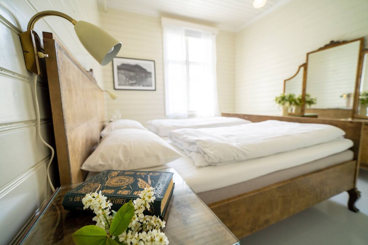 A neatly made bed with white linens, a bedside lamp, and a book with flowers on a bedside table.