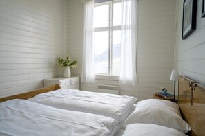 A bedroom with two beds, a nightstand with a lamp, a window with curtains, and a view of mountains. - Sommerhotellet (Vang)