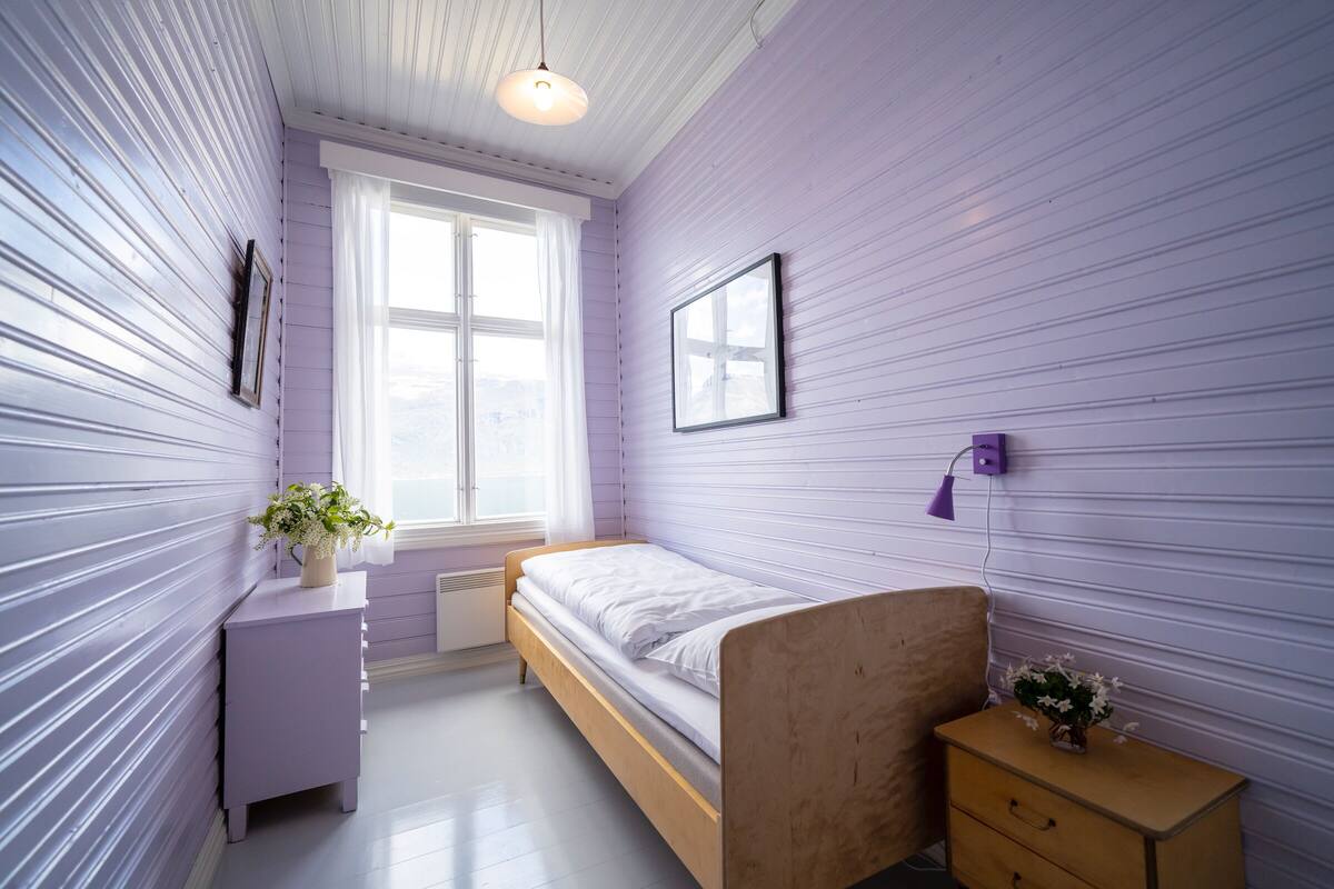 A bedroom with a wooden bed, a small bedside table, a nightstand, a window with curtains, and a wall-mounted lamp.
