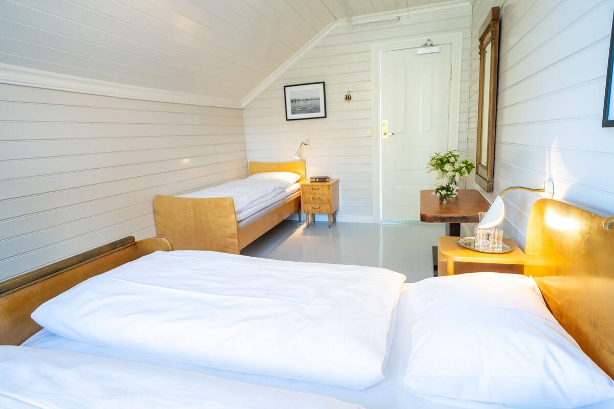 A hotel room with two beds, a bedside table with a lamp, a small bedside table with a vase of flowers, and a framed picture on the wall.