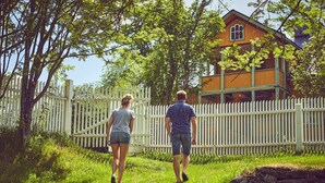 Two people walking on a grassy path with a white picket fence and a wooden house in the background. - Sommerhotellet (Vang)