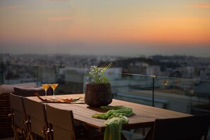 Una taula de menjador amb una planta en test, dues copes de martini i un drap verd a un balcó amb vista a un paisatge urbà al capvespre.