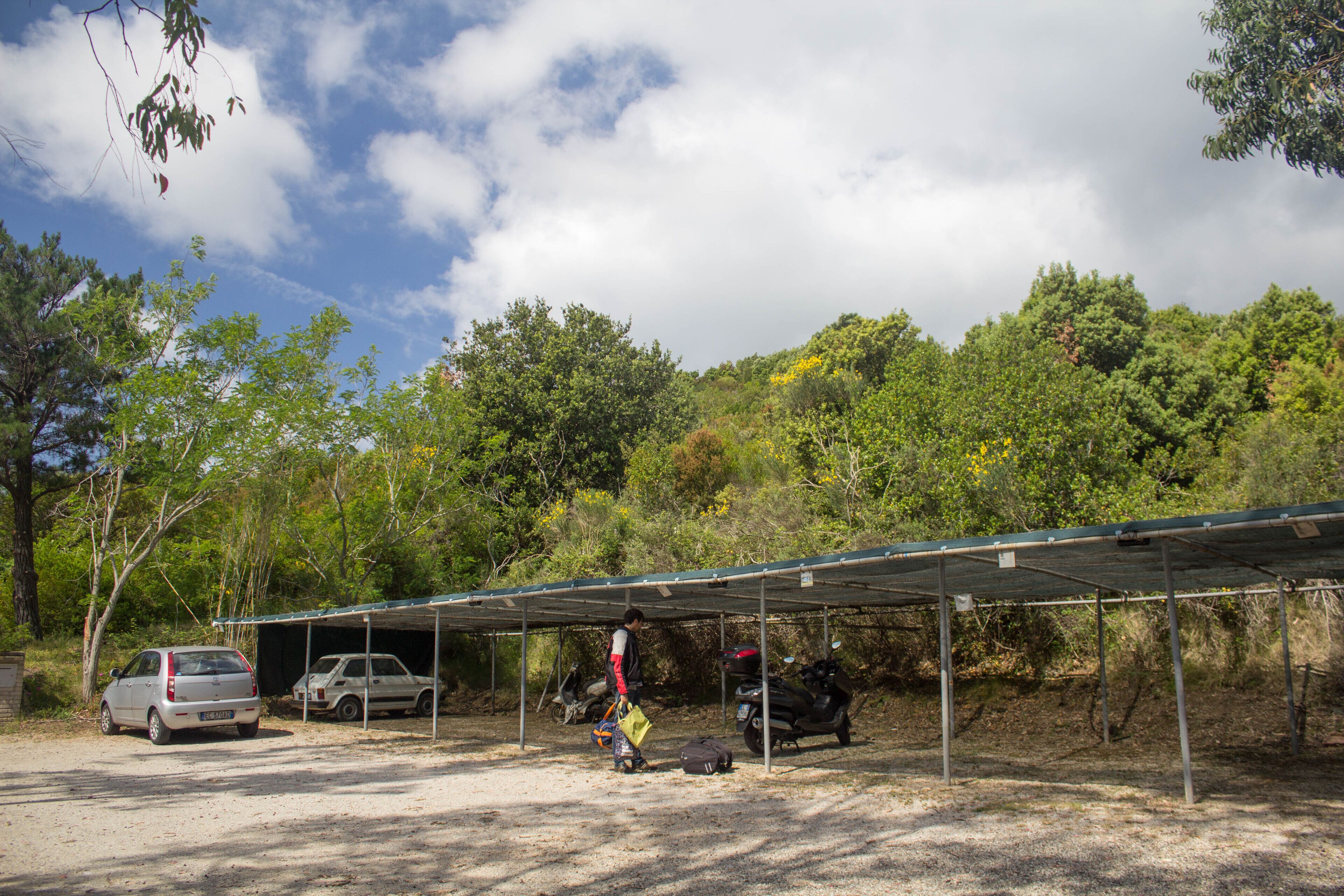 A covered parking area with a car and a motorcycle, surrounded by trees and a clear sky.
