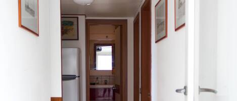 Un couloir étroit, avec un réfrigérateur sur la gauche, un espace avec un évier au milieu et une porte sur la droite.
