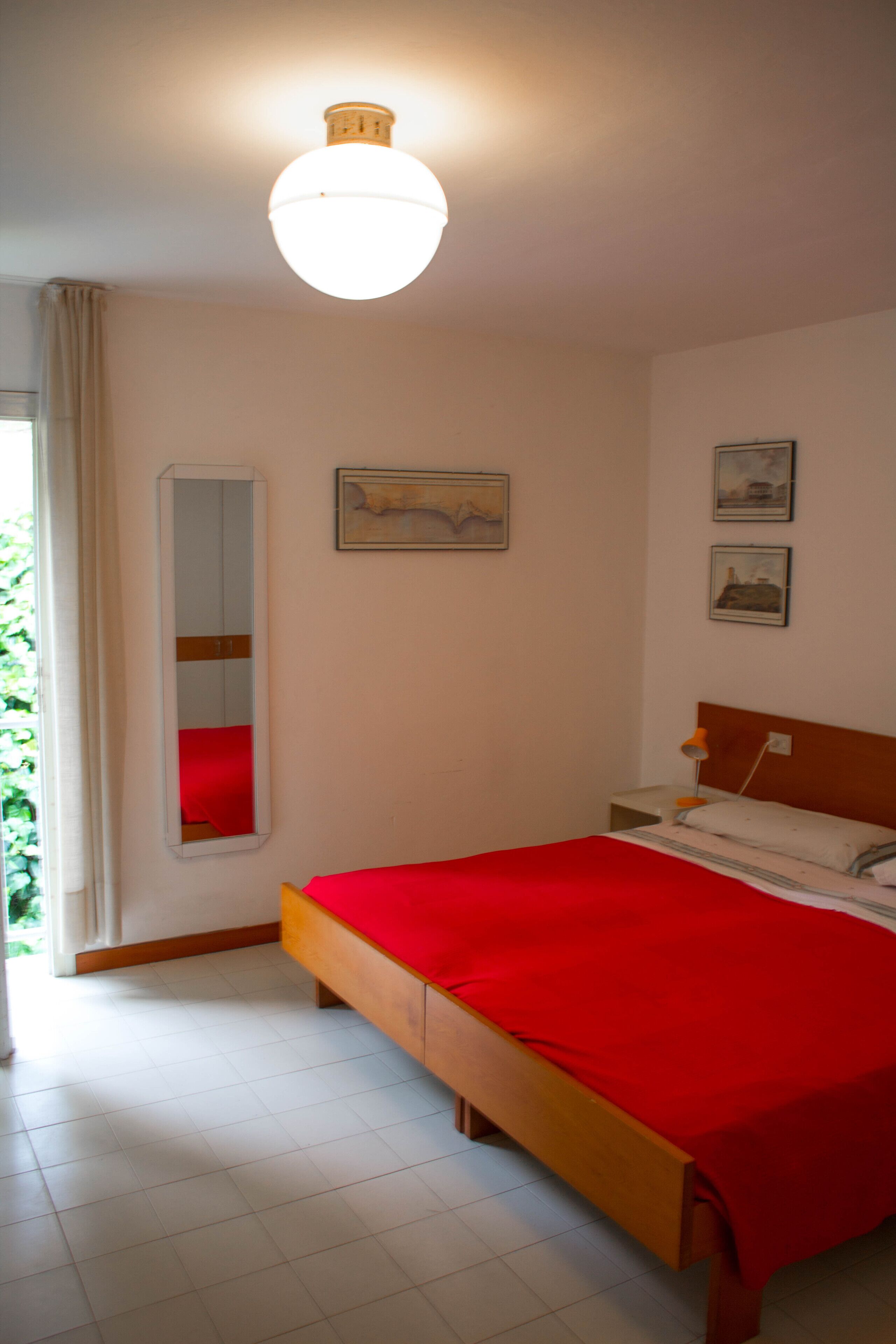 Una camera da letto con un letto di legno, una coperta rossa, un comodino con una lampada e uno specchio appeso al muro.