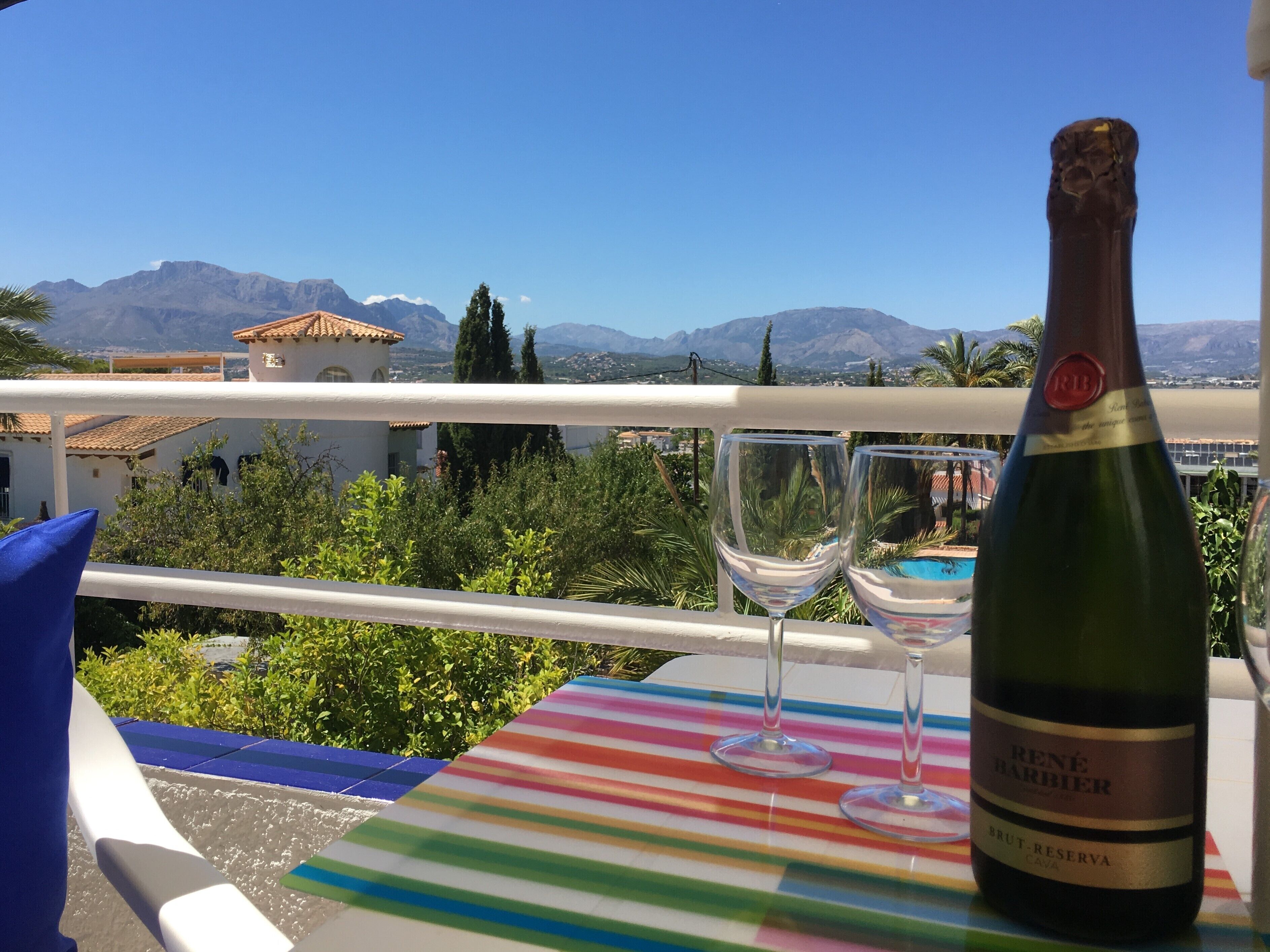 Un balcón con una botella de vino y dos copas sobre un mantel a rayas, con vistas a un paisaje montañoso.