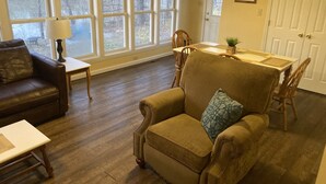 A living room with a brown leather sofa, a tan armchair with a patterned pillow, a wooden dining table, and a window with a view of trees.