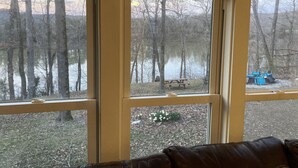 A room with three windows overlooking a wooded area with a lake and picnic tables.