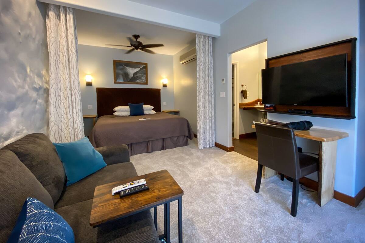 A hotel room with a bed, a TV, a desk, a chair, a sofa, and a remote control on a small table.