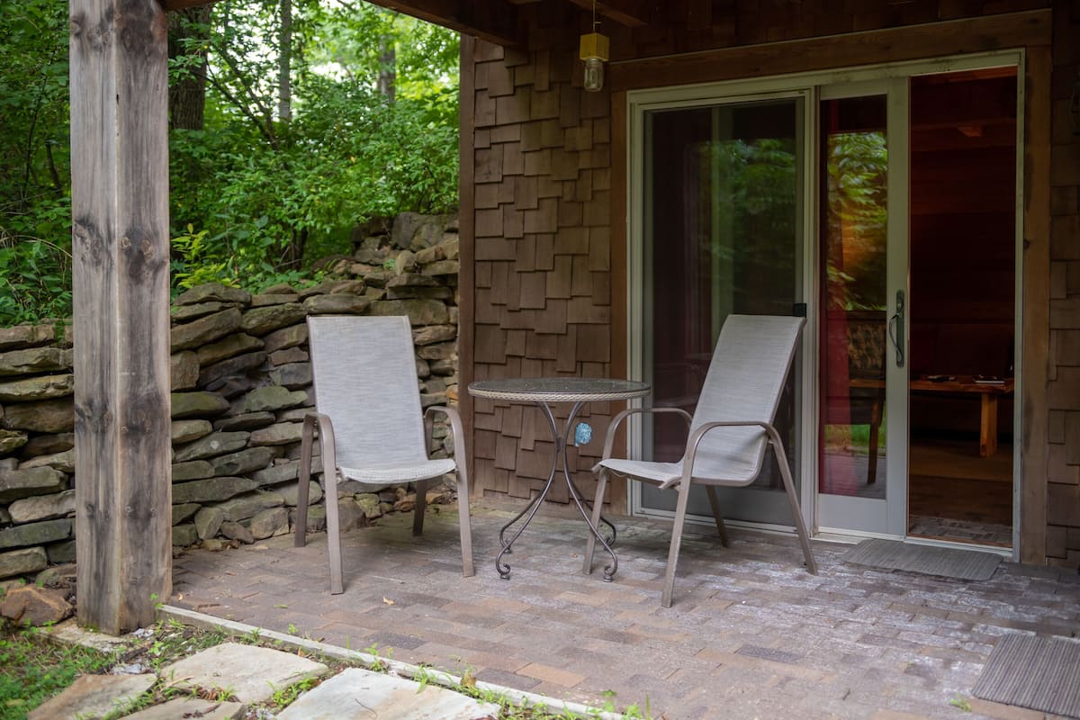 An outdoor patio with two metal chairs and a small round table, set against a stone wall and wooden deck.