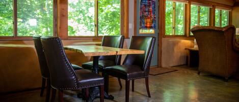 A dining area with a wooden table and chairs, large windows with a view of greenery, and a hanging light fixture.